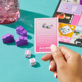 Hand holding a Barbie-themed card game card with dice and game pieces on a teal surface