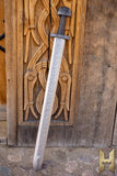 Decorative sword with intricate blade design leaning against a wooden wall with carvings.