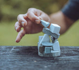 A hand carefully balancing a small stack of geometric stone blocks in white, and gray colors on a dark wooden table with a blurred green outdoor background.