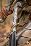 Decorative sword with ornate handle held by a person in armor, blurred natural background