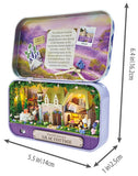 A decorative metal tin shown open, featuring a miniature village diorama with cottages, trees, flowers, and a lavender field scene on the inside lid. Black dimension arrows and text indicate measurements of approximately 6.