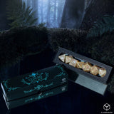 A boxed set of polyhedral gaming dice displayed in a dark forest-themed setting. The set includes a black decorative box with turquoise line art and branding, alongside an open black tray holding multiple gold-toned polyhedral dice.