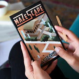 Mazescape board game box held by hands with a pen, on a blurred background.