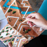 Hand holding a wooden stick over a board game map with paths and trees.