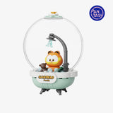 A toy bath scene under a clear dome with a cartoon orange cat character sitting in a bathtub-like base, a shower arm with water spray, and a visible PAN TASU logo in the top right on a light gray background.