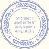 A circular decorative plate-style graphic with a light blue border, leafy wreath motif, and centered text reading, 'SWITCH HANDS: IF YOU ARE A LEFTY, EAT RIGHTY. IF YOU'RE A RIGHTY, EAT LEFTY.