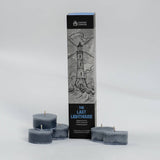 A tall black and gray candle box labeled 'The Last Lighthouse' stands upright on a light gray studio background, with four matching dark tealight candles arranged in front of it.
