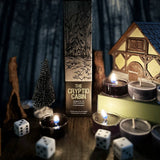 A boxed game product labeled 'THE CRYPTID CABIN' stands upright on a dark tabletop surrounded by candles, dice, a small tree, driftwood, and a house-shaped decoration in a moody styled setting.