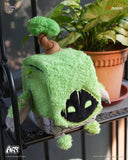 Green plush toy with black face on a shelf with plants in the background