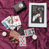 A Wack This Deck card deck set arranged on a purple quilted blanket with candles and stones. The set includes a black box with a skull graphic, a printed card back reading 'Wreck This Deck' by Becky Antoon, several illustrated playing cards, and small decorative stones and candles.