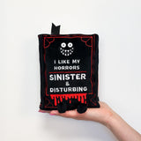 Small book-shaped plush with a spooky design and text held by a hand on a white background