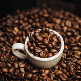 White cup filled with coffee beans surrounded by roasted coffee beans on a dark background