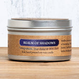 Candle in a metal tin labeled 'Realm of Shadows' on a wooden surface.