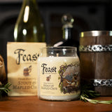 A candle in a clear glass jar labeled 'Feast' by Feast Wanderers, styled on a wooden surface with a bottle, barrel, and paper bag in the background.