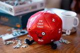 Red die-shaped plush toy with text on a table with puzzle pieces and a mug.