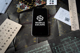 Phone displaying 'Mobile Escape' logo on a table with game cards and pieces.