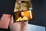 Hand holding a smartphone displaying a video call with a person, with a wooden table and papers in the background.