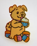 Enamel pin of a cartoon dog holding a cupcake on a white background