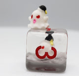 A small white ghost figurine with red eyes and pink cheeks sits on top of a clear square glass block containing a larger matching ghost figure with a red heart and black-and-yellow hat, photographed against a light gray background.