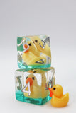 Two translucent acrylic polyhedral dice cubes stacked vertically, each containing a small yellow rubber duck inside, with a separate small yellow rubber duck placed beside them on a light gray background.