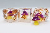 Three translucent polyhedral dice with embedded purple mushrooms and gold numbers, displayed on a light gray background.