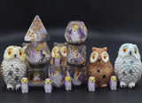 A set of owl-themed tabletop gaming accessories, including four large owl figurines in white, brown, and pale blue-gray, and multiple translucent polyhedral dice with gold numbers and purple glitter, arranged on a dark surface against a dark background.