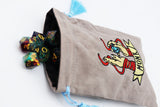 A beige drawstring pouch with a blue tassel and an embroidered tattoo-style graphic of hands holding a gem, with the word 'MONK' on a banner. Several multicolored polyhedral dice are spilling out of the pouch against a white background.