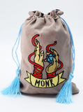 A taupe drawstring pouch with a blue tassel and an embroidered graphic of two monk hands holding a blue gem, with a banner reading 'MONK'.