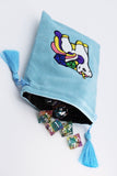 A light blue velvet drawstring bag with a colorful embroidered unicorn design on the front. The bag is shown open with a black interior, light blue tassels on both sides, and several translucent multicolored polyhedral gaming dice spilling out.