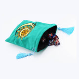 A teal green velvet drawstring pouch with a gold crescent moon and sun design on the front, shown spilling out polyhedral dice against a white background.