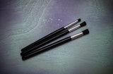 Three black makeup brushes with silver ferrules on a textured gray surface
