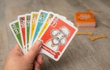 Hand holding a set of dog-themed playing cards on a wooden surface with a box and bone-shaped objects in the background.