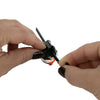 Close-up of a person's hands demonstrating how to use a small black multi-tool or keychain tool with a small orange accent and a silver ring, against a plain white background.