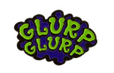 Enamel pin with 'GLURP GLURP' text in green and purple on a white background