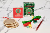 Wooden Christmas ornaments cross stitch kit laid out on a white marble surface, including a red and green instruction booklet, a green pattern box, two round wooden peg frames, colorful thread bundles, a red pencil, and a red ribbon.