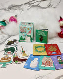 A Christmas-themed card game set displayed on a white marble surface with decorative ornaments and white faux fur. The set includes a mint green box labeled 'Merry Mix 'N Match', a green booklet, and several illustrated cards and cutout pieces showing holiday scenes and characters.