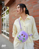 Woman holding a purple plush bag with a face design against a brick wall.
