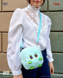 Person wearing a white blouse with lace details, holding a fluffy turquoise bag against a brick wall.