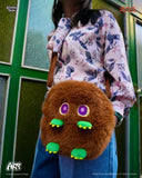 Person wearing a floral shirt with a brown plush bag featuring green feet and purple eyes.
