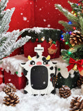 A cow-shaped blackboard with a cross on its forehead, styled in a festive Christmas scene with snow, pinecones, a decorated tree, and red gift boxes. The brand name 'Globion' is visible on the crosspiece.