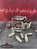 Metal dice holder with dice and a red die on a dark surface with Elvish script in the background.