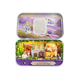 A miniature dollhouse set inside a purple tin box labeled 'Peaceful Getaways LILAC COTTAGE,' shown open against a white background. The lower compartment features a detailed cottage scene with small houses, flowers, greenery, and tiny furniture pieces.
