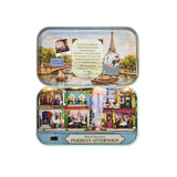 A Paris-themed diorama miniature set in a decorative tin box, shown open to reveal a detailed city scene with Eiffel Tower, boats on the river, and colorful street buildings labeled Hotel & Restaurant and Cafe.