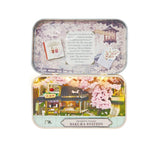 A rectangular metal tin shown open, revealing a miniature diorama model of a Japanese sakura station with pink cherry blossoms, small buildings, and warm interior lights. The top lid has printed cherry-blossom artwork and a small card insert.