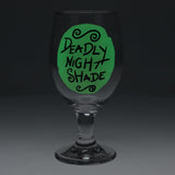 A clear stemmed wine glass with a green circular graphic on the bowl featuring the hand-lettered text 'DEADLY NIGHT SHADE' in black, shown against a dark gray studio background.
