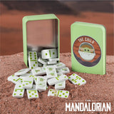 White ceramic dominoes with green dot details are shown spilled from a light green metal tin next to its closed tin, which features a Star Wars The Child logo, set on a reddish-brown sandy surface.