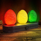 Three textured egg-shaped lamps in red, yellow, and green sit on a dark carved wooden base with a triskelion emblem, displayed on a rustic wooden table against a dark background.
