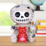 Mummy figurine with a red teddy bear toy on a wooden surface
