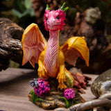 A small fantasy dragon figurine with pink and yellow coloring, standing on a rocky base with flowers, photographed in a styled natural-looking setting with blurred wood and stones.