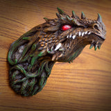 A detailed fantasy dragon head wall sculpture with textured scales, green accents, and a red eye, displayed on a wooden surface.